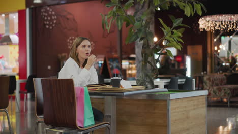 dama sentada sola en una mesa en una zona de comedor de un centro comercial, con una bebida, papas fritas y un libro abierto, capturada en un momento acogedor y relajado con un fondo vibrante con atmósfera de centro comercial