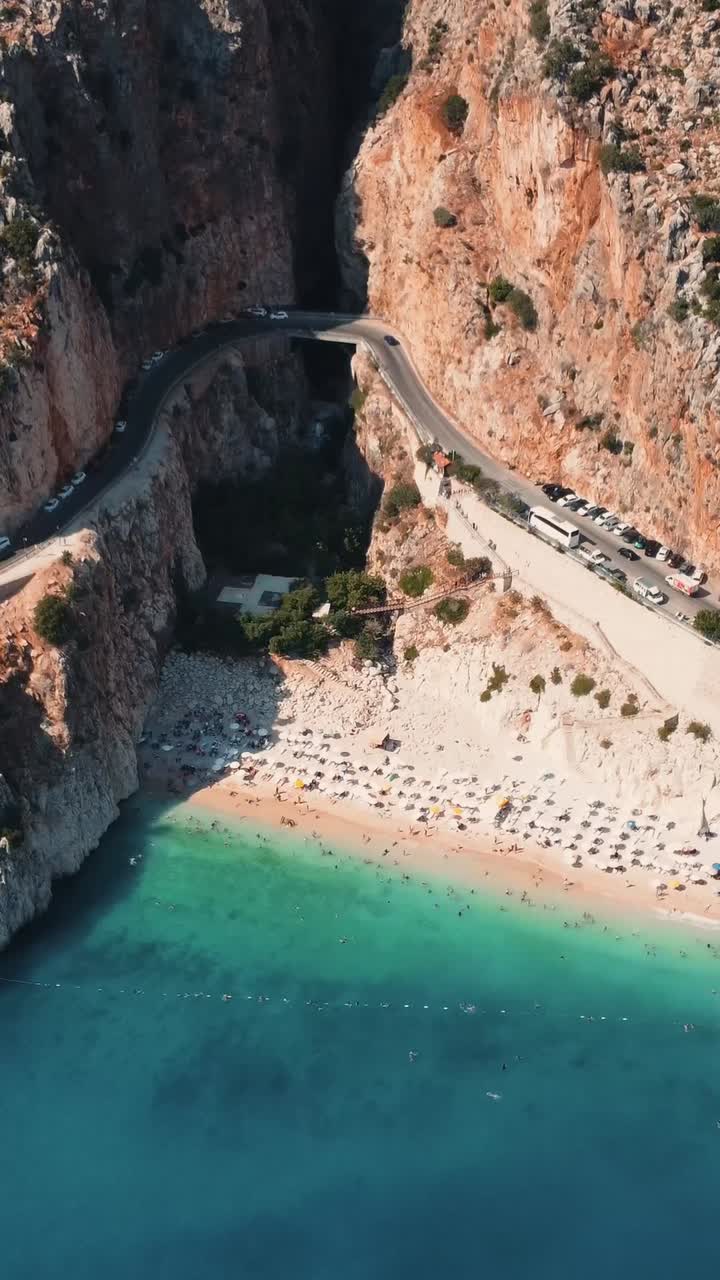 Aerial drone social media style video showing curved cliffside beach with sunbeds and swimmers beside parked tour buses on winding coastal road at Kaputas Beach in Kas Antalya Province