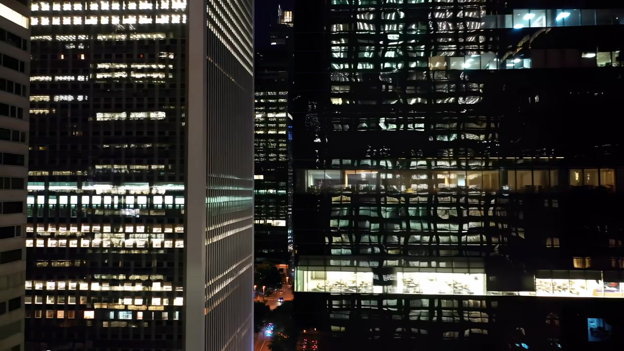 Vista ravvicinata di due facciate di grattacieli adiacenti di notte, con illuminazione delle finestre mutevole, superfici di vetro riflettenti, motivi a griglia verticale e un'atmosfera cupa di città aziendale, con uffici illuminati nella notte