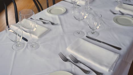 Dining table set for a wedding or corporate event at fine dining restaurant ceramic plates forks knives cloth napkins on white tablecloth on table steady slow motion medium-wide panning upward