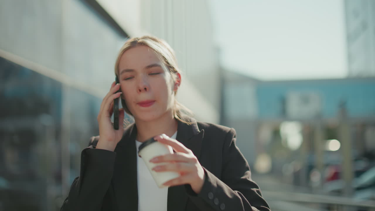 White woman on call drinking coffee while walking by office building with cup in hand, dressed in professional attire, with blurred parked cars in background and sunlight illuminating her