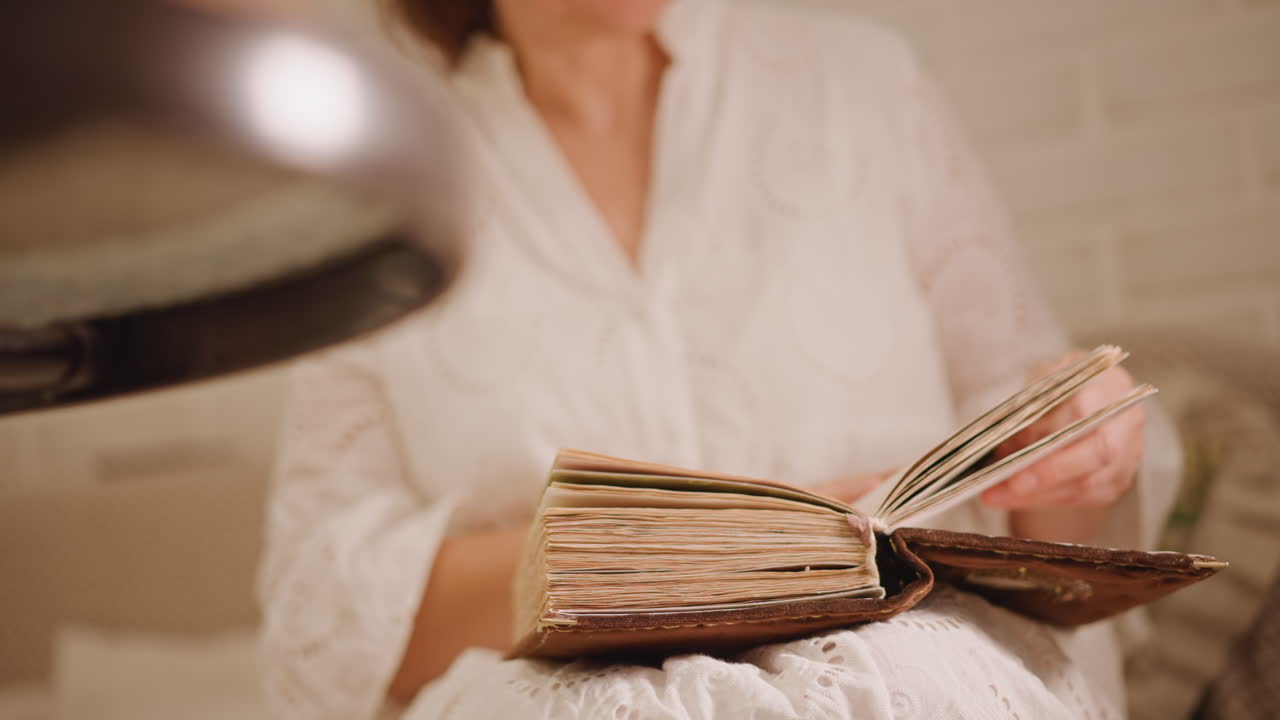 Donna che sfoglia un antico quaderno di ricette accanto a una lampada calda, pagine che si aprono in una luce soffusa, abito ricamato e forno visibile a lato, torta e teglia in secondo piano sfocato, sfogliare le pagine in modo tattile, lento e cinematografico.