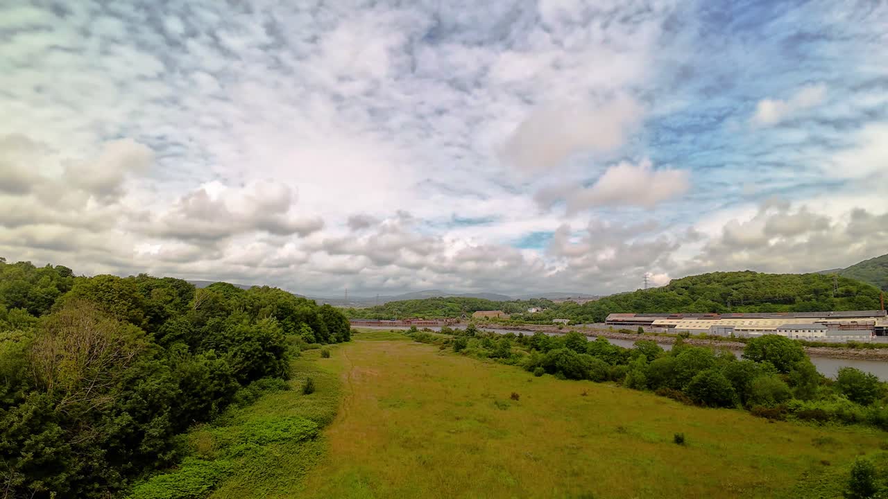 Wide Angle Timelapse of Long Grass Blowing Patterns in the Wind with Blustery Windy Trees and Clouds Passing Quick in the Sky with Warehouses in Distance near River Neath in South Wales.