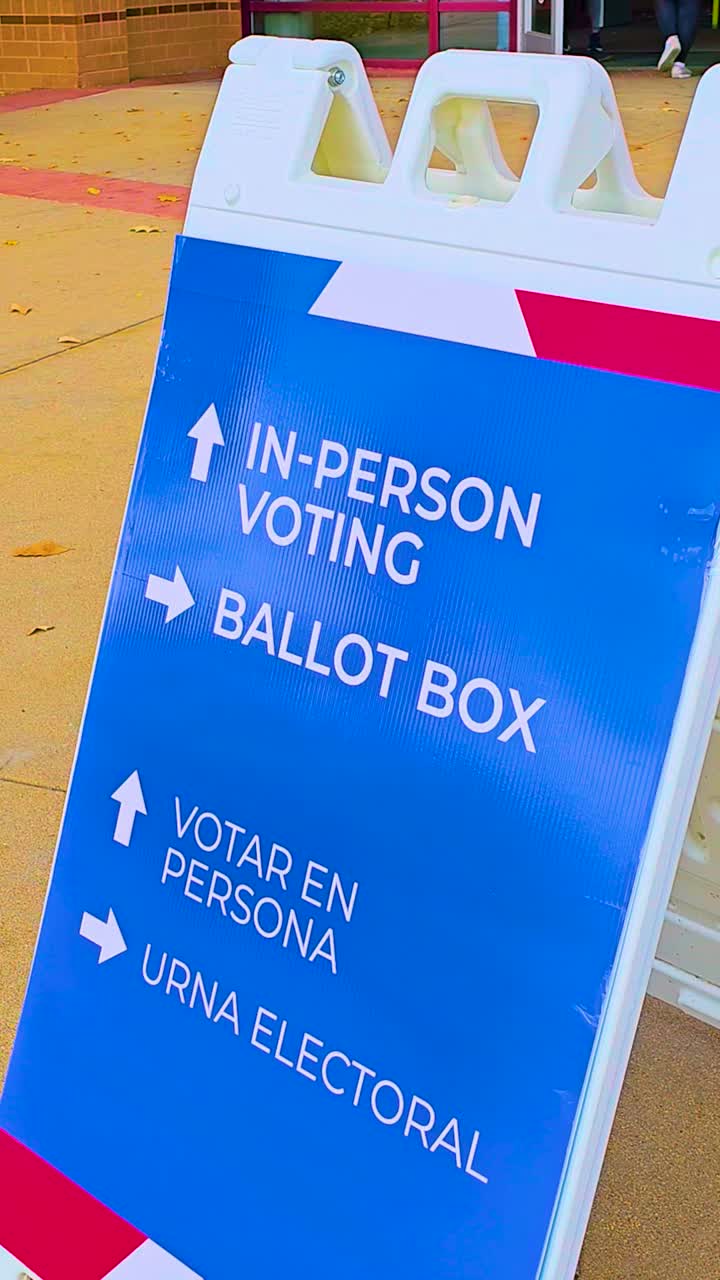 Pedestrians move past while a ballot drop-off sign with bold arrows shows Spanish text Votar en Persona and Urna Electoral, highlighting civic responsibility, safety, democracy, freedom. Vertical