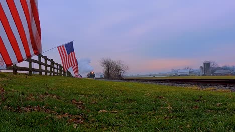 a low angle view of a line of gently waving american flags on a fence by farmlands as a steam passenger train blowing smoke approaches during the golden hour-1