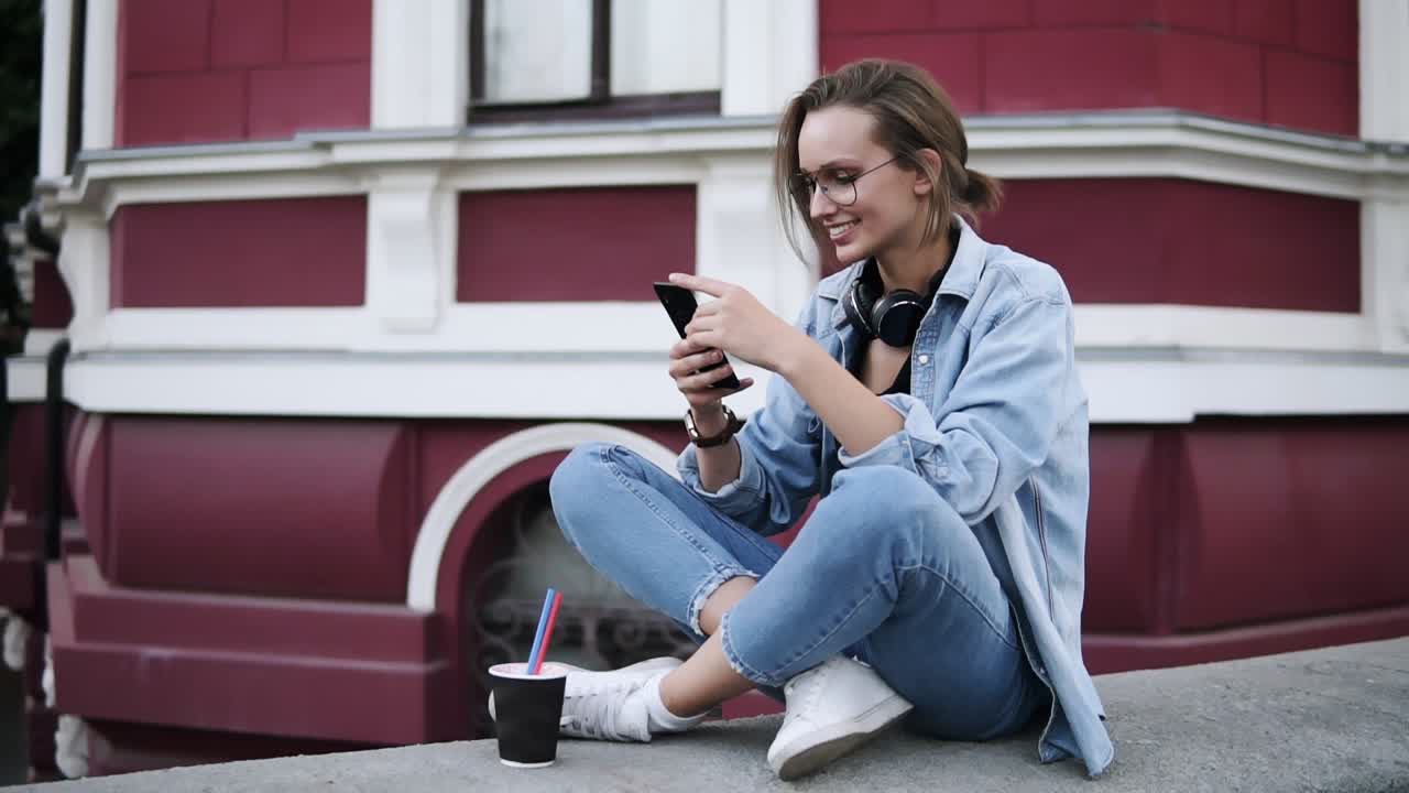 A fashionable girl in glasses sits on the street on a parapet, concentrates on her mobile phone. Smiling, communication