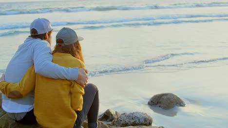 Rückansicht-Eines-Jungen-Kaukasischen-Paares,-Das-Auf-Einem-Felsen-Sitzt-Und-Am-Strand-Auf-Das-Meer-Blickt,-4k