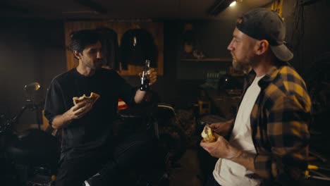 Mechanic-guy-in-a-plaid-shirt-and-his-friend-eat-a-burger-and-drink-soda-during-their-lunch-in-a-workshop-studio