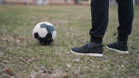 close-up of a person in a black outfit standing near a soccer ball on a grassy field, with the blurred view of building in the background