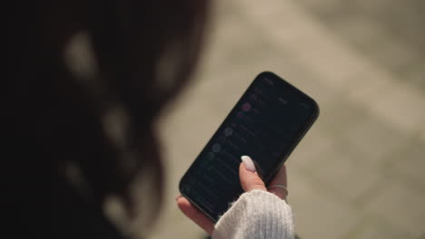 close-up of woman with short hair in black coat and white sweater scrolling on phone with focus on hand holding phone with visible manicure, gentle outdoor light, and blurry background