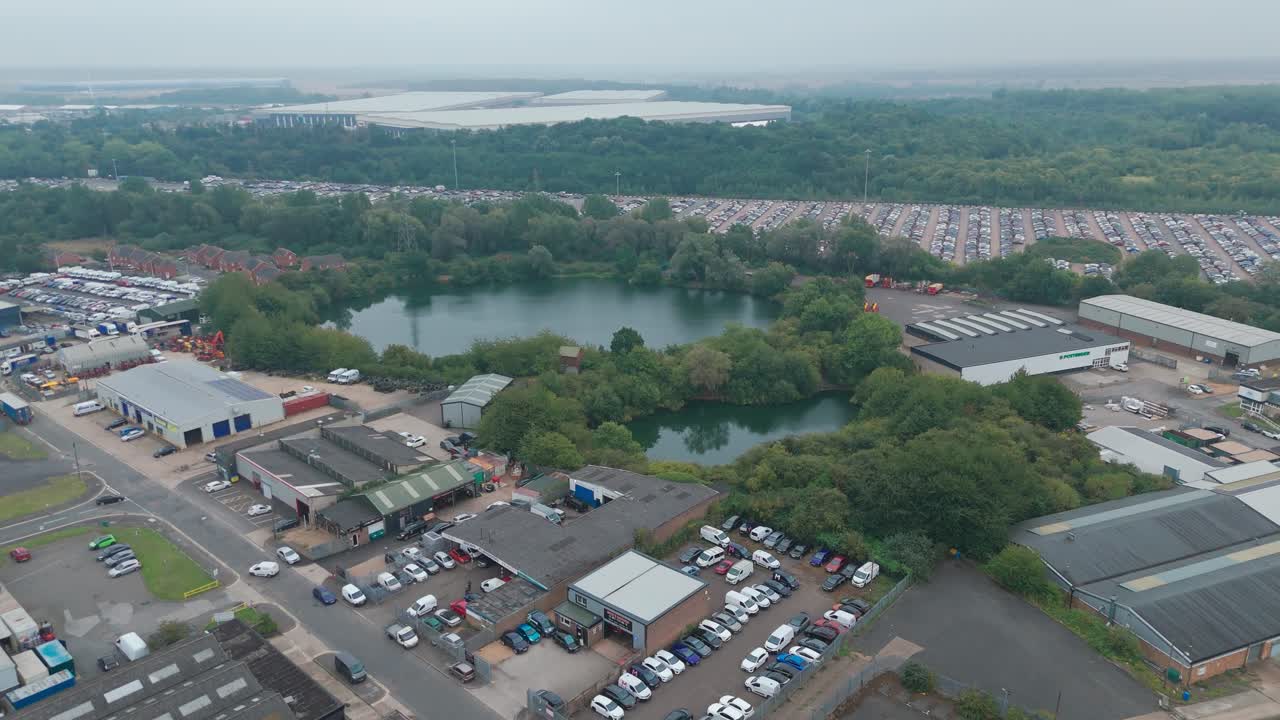 Aerial shot of an industrial zone featuring buildings, parked cars, and a water body under a cloudy sky. The area includes warehouses, roads, and distribution centres