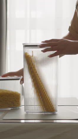 housewife examines packaged raw spaghetti slow motion. woman stacks containers on table with bouquet evaluating convenience of using plastic kitchen utensils