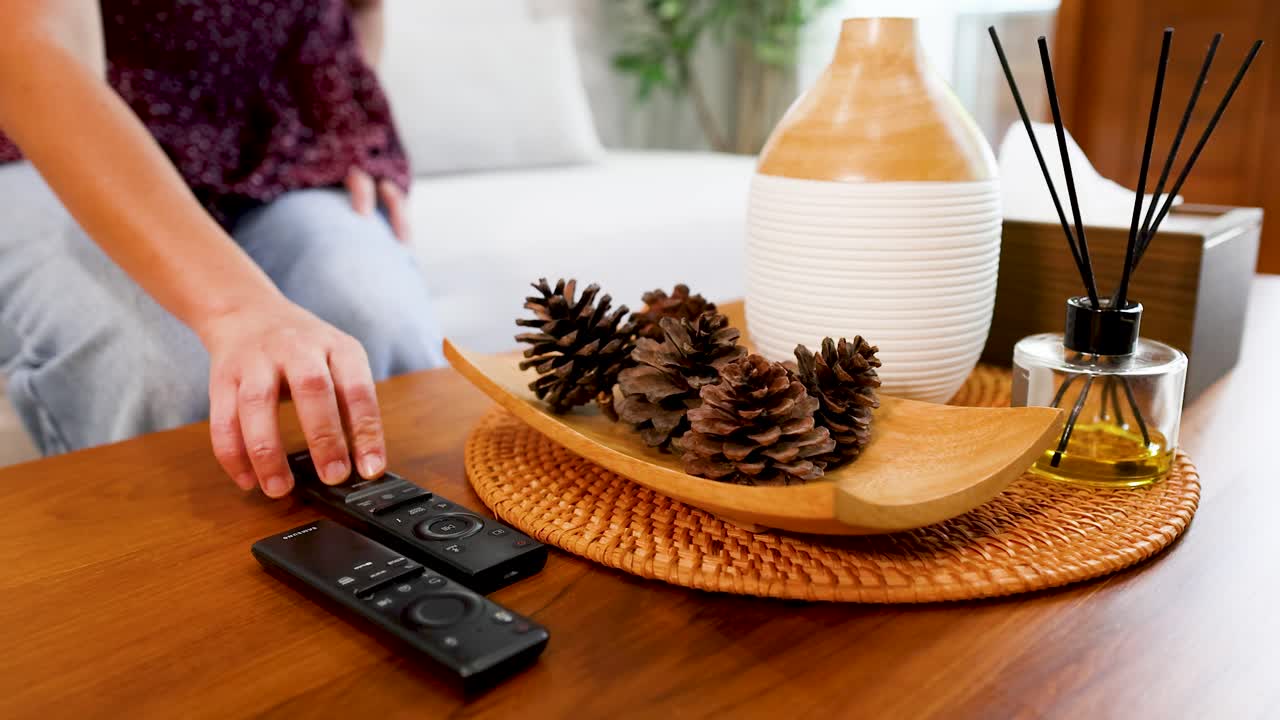 Une femme tend la main vers une télécommande noire posée sur une table en bois avec des objets décoratifs.