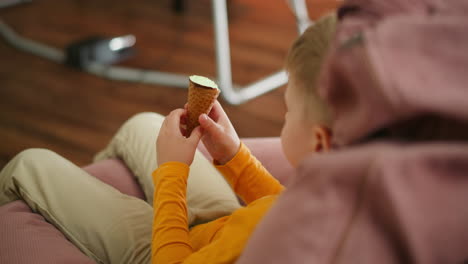 aerial view of young boy in orange top relaxing barefoot on pink beanbag while eating ice cream cone, casually playing with legs, with blurred metal stand and wooden floor visible in soft background