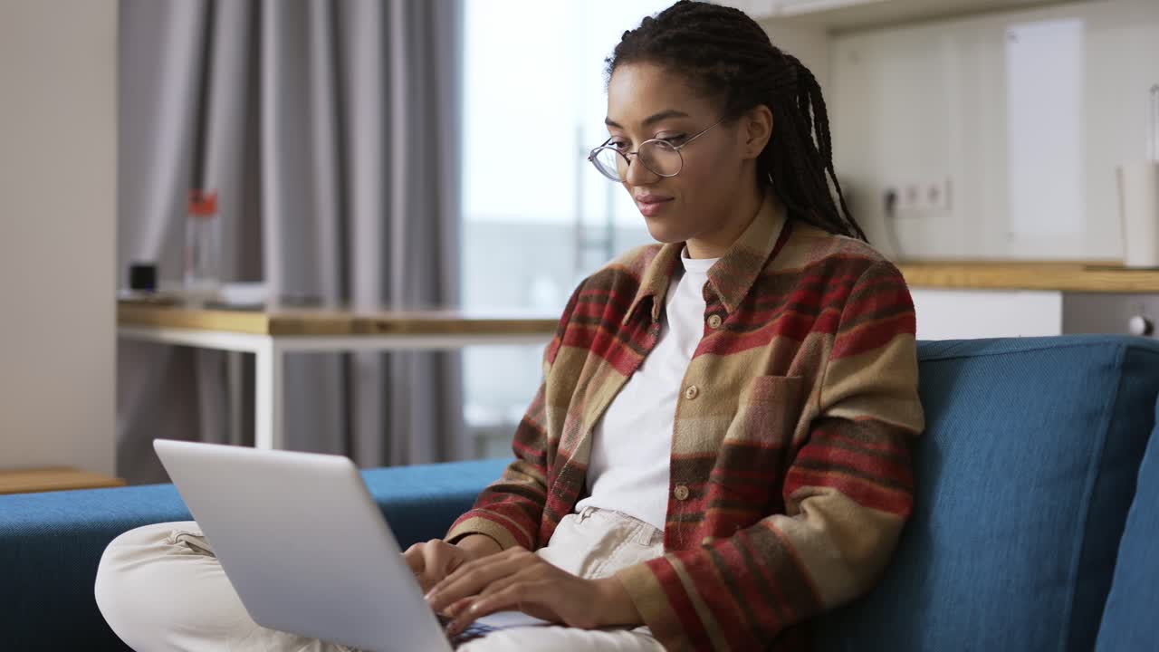Woman with dreadlocks is working on new project and using laptop on couch in apartment room, slow motion