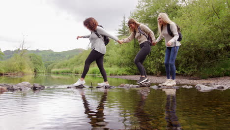 three young adult women hold hands while carefully crossing a stream on stones during a hike