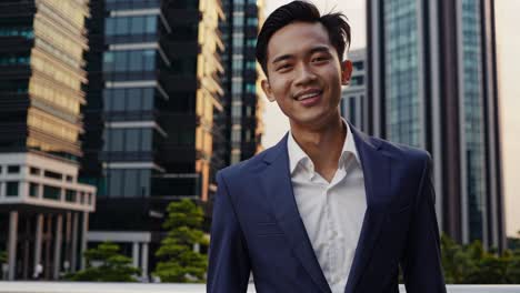 professional asian manager wearing tailored suit, standing confidently with urban office skyline during golden hour, representing career success and corporate ambition