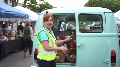 a woman in a reflective vest joyfully reveals a van filled with fresh bread and flowers at a bustling outdoor market, surrounded by colorful stalls and lively shoppers