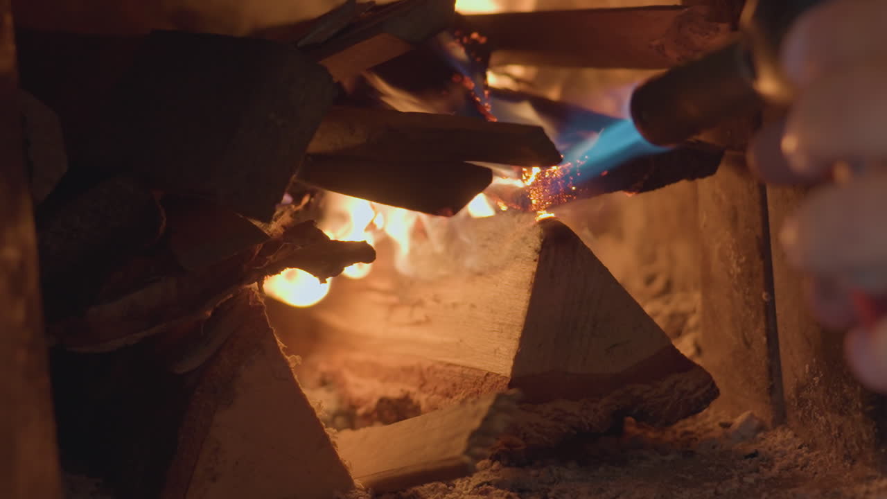 Close up of person using blue gas torch to ignite stacked firewood inside dark fireplace, wood catches flame slowly, emitting glowing embers and flickering light in warm cozy indoor environment