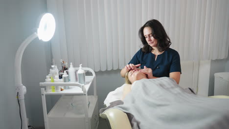 facialist massaging under eye area of relaxed participant on massage bed with creamy product, gentle moisturizing strokes smoothing delicate skin around eyes for rejuvenation in serene treatment