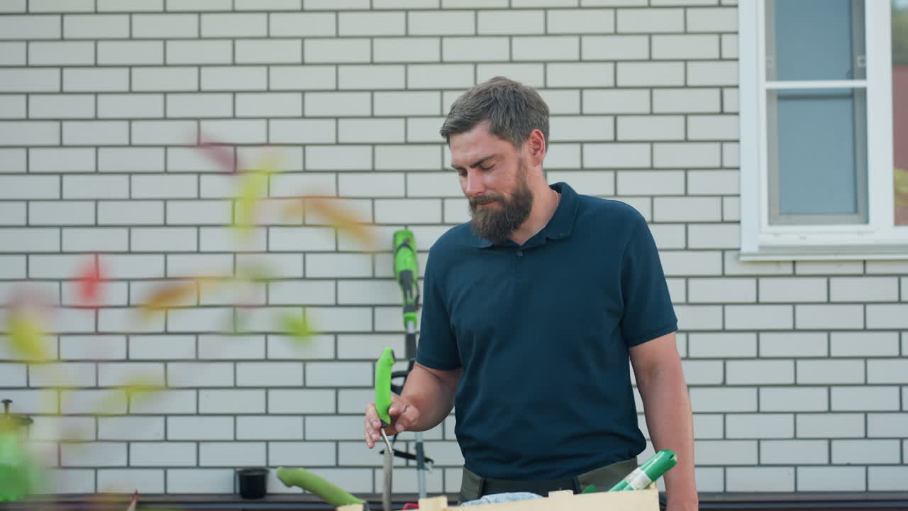 Bearded man in dark shirt standing outdoors near white brick wall drops trowel with green handle into wooden crate filled with garden tools during daytime work under clear sky