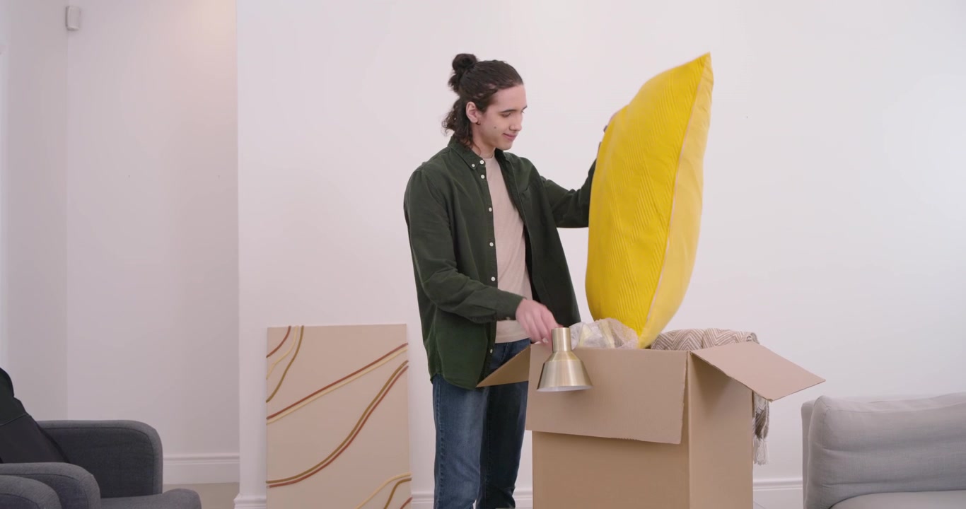 African American man opening moving box, lifting gold lamp and yellow pillow, preparing living room