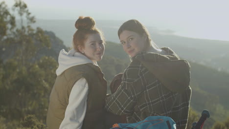 two young female hikers sitting at cliff edge and talking, then turning to the camera and smiling