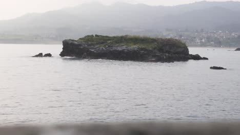 Small-island-in-the-middle-of-the-ocean,-mountains-at-the-background,-Asturias,-Spain,-static-shot