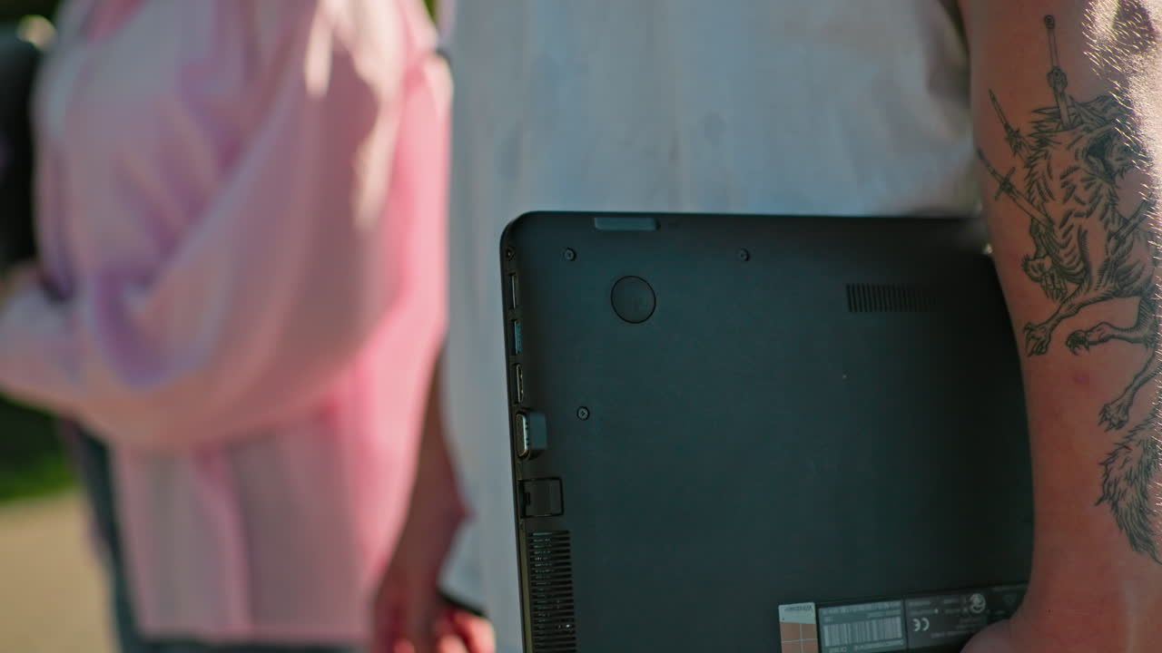 핑크 을 입은 사람과 함께 는 동안 노트북을 들고 있는 문신한 팔의 클로즈업, 밝은 빛 아래 문신 예술의 캐주얼 상호 작용과 세부 사항을 포착합니다.