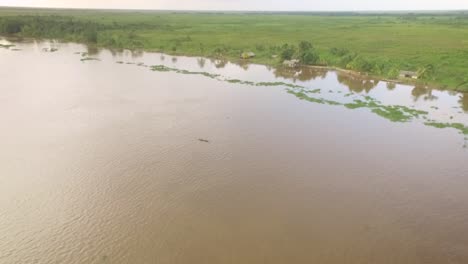 Vista-Aérea-De-Una-Pequeña-Canoa-Indígena-Que-Navega-Por-El-Río-Orinoco-Con-Pequeñas-Casas-Indígenas-En-La-Orilla