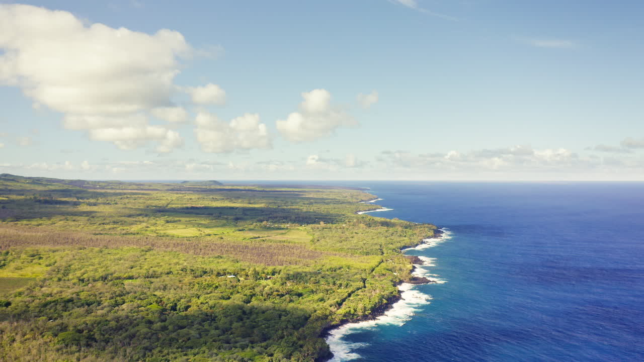 The coastline of Hawaii’s Big Island unfolds beneath scattered clouds, where emerald-green forest meets the endless blue of the Pacific in a stunning contrast of color and texture