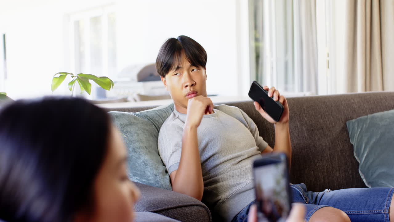 Yawning and holding smartphone, Asian man sitting on couch while woman takes photo