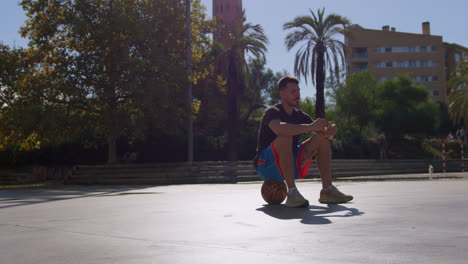 general shot of a young caucasian man sitting on top of a basket ball, relaxed eating an energy bar on a street basketball court in barcelona, spain