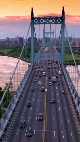 the triborough bridge with moving cars against river and city panorama. manhattan skyscrapers at the backdrop of orange sky. vertical video