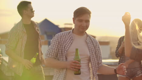 The-men-is-dancing-on-the-roof-with-his-friends-who-drinks-beer-on-the-party.-He-smiles-and-enjoys-the-time-in-shorts-and-a-blule-plaid-shirts-in-summer-evening.