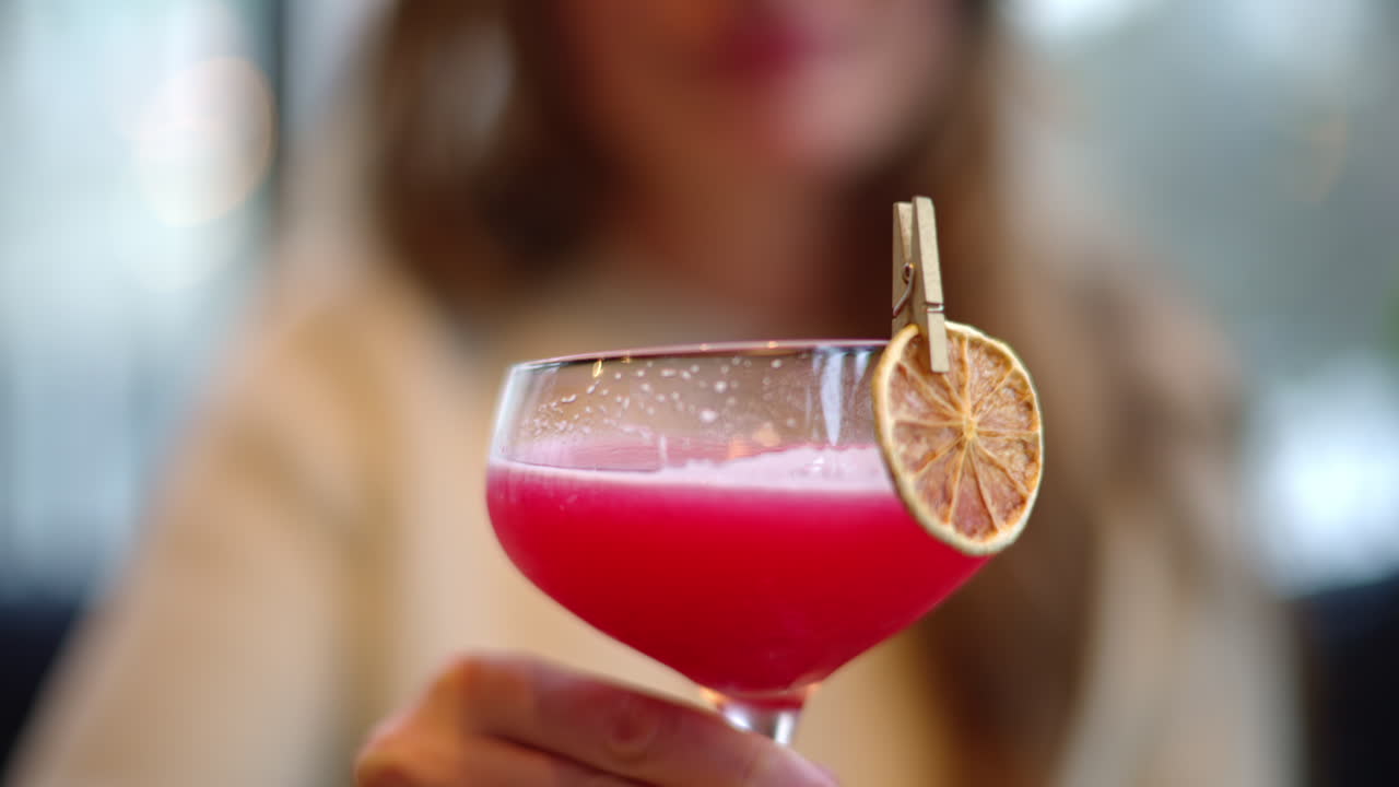 Seorang wanita minum minuman koktail merah dengan dekorasi berupa jeruk kering