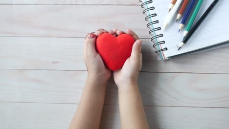 child's hands holding a red heart on a wooden table with stationery