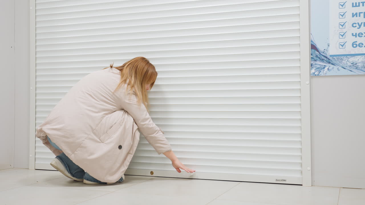 Laundry manager closes white rolling shutter at shop entrance, arms raised, dim interior with washing machine visible, corridor signage, casual jacket and jeans, clean indoor service scene