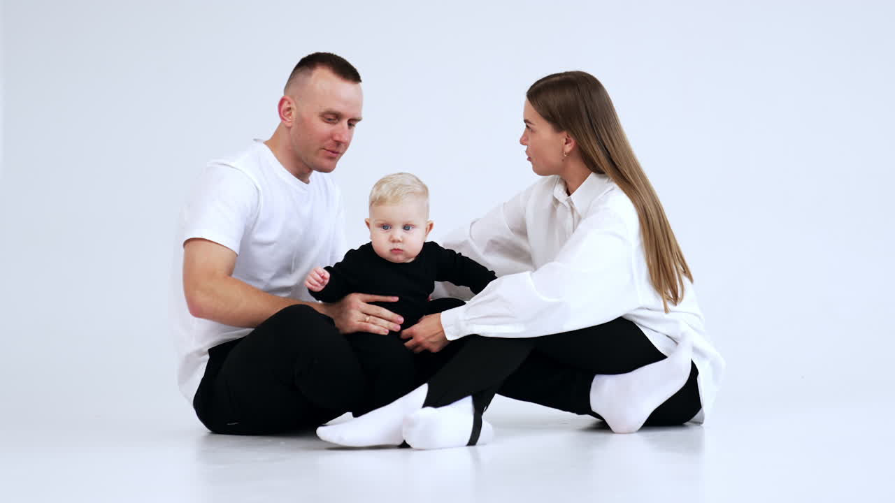 Smiling woman and man sit on the floor. Loving parents try to hold their active baby. White backdrop.