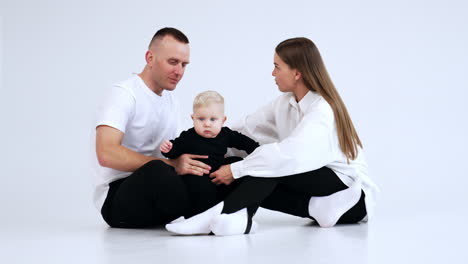 smiling woman and man sit on the floor. loving parents try to hold their active baby. white backdrop.