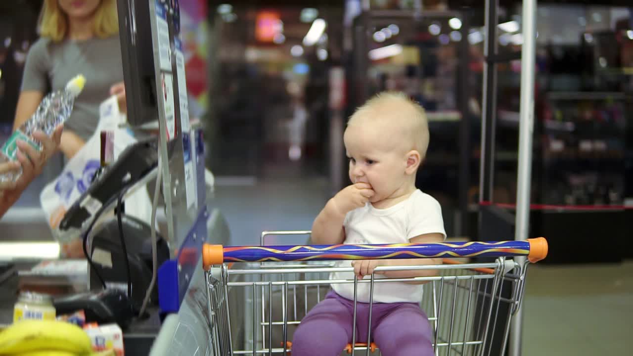 madre joven sacando de los productos del carrito en la cinta transportadora en el supermercado mientras su bebé lindo está tomando plátano