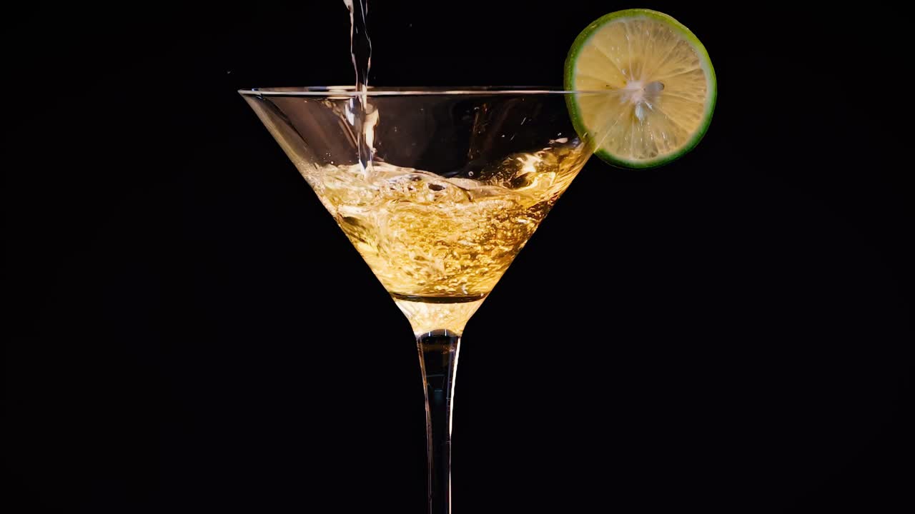 A vibrant golden liquid is poured into a martini glass with a lime slice garnish, creating dynamic splashes.