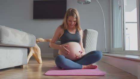 pregnant woman wearing fitness clothing on exercise mat at home touching stomach