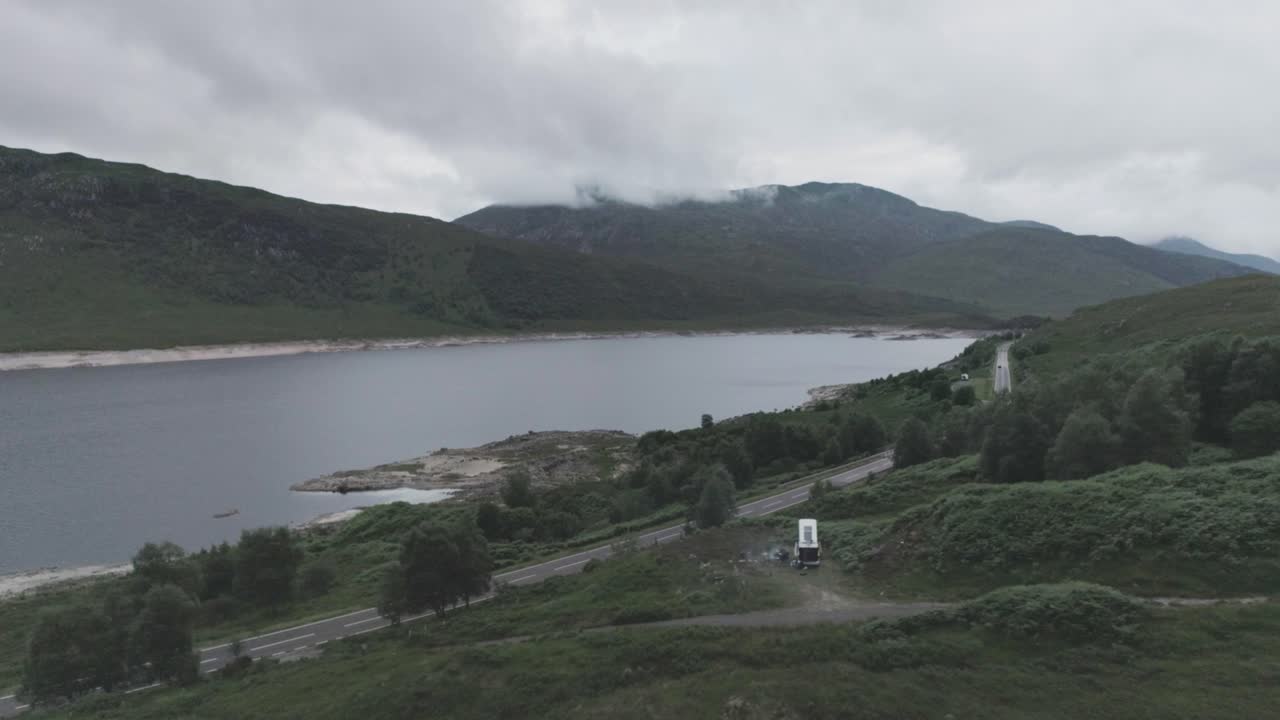Super wide pull back drone shot of Loch Beag showing a campervan wild camping, BBQ smoke, coastal road and cloudy mountains in the background in the Scottish Highlands