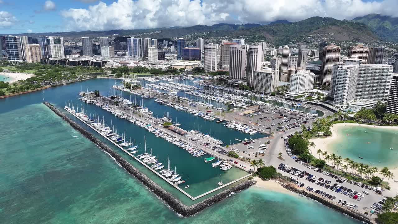 Aerial drone footage of Ala Wai Boat Harbor in Honolulu, Oahu, Hawaii, showing boats, tropical waters, palm trees, and the scenic Waikiki coastline with hotels and city skyline in the background