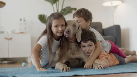 niños felices tirados en el suelo y abrazando a un perro encantador en casa y mirando la cámara