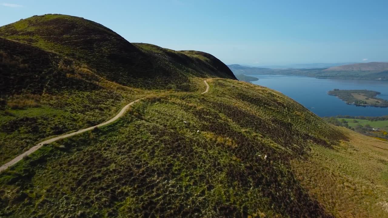 Aerial view above winding mountain road during a beautiful sunny day and blue sky, rural scenery. Majestic rolling hillside, blue freshwater lake with a green island is behind, Scottish loch landscape