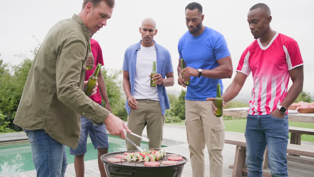 Grilling man in olive jacket turning skewers on charcoal grill, deck by pool, group holding beers
