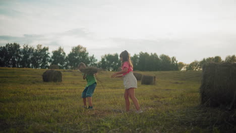 excited siblings joyfully toss hay in open countryside field at dusk, laughter fills air as they play freely, surrounded by nature, golden sunlight casts a warm glow