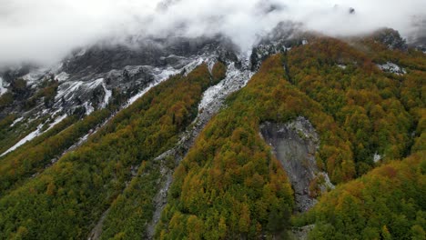 Picos-Montañosos-Cubiertos-De-Nieve-Y-Escondidos-En-La-Niebla-Y-La-Niebla-En-Los-Alpes,-Sobre-El-Bosque-Colorido-Con-árboles-Altos